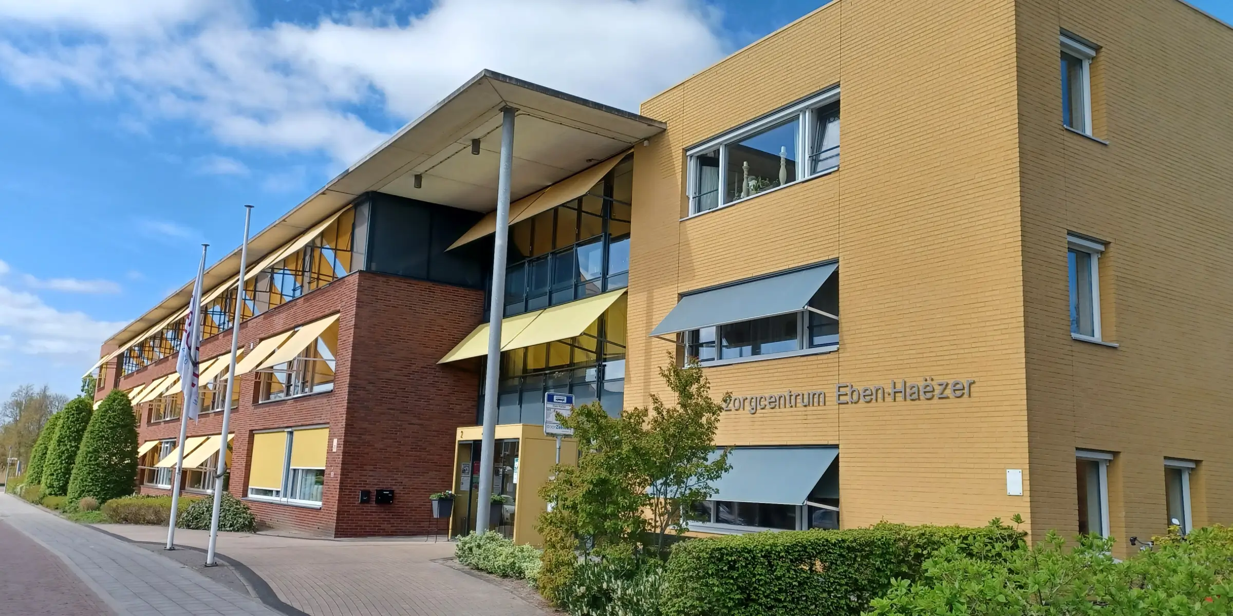 Außenansicht des Pflegezentrums Eben‑Haëzer mit gelber Klinkerfassade, großen Fenstern, Markisen und grünen Sträuchern vor blauem Himmel.